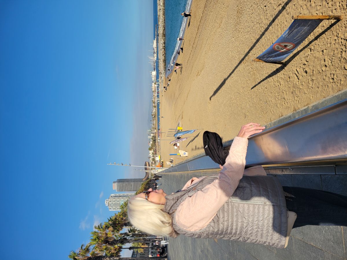 Shirley leaning on the railing of the Barceloneta promenade, looking toward the sandy beach and the sea
