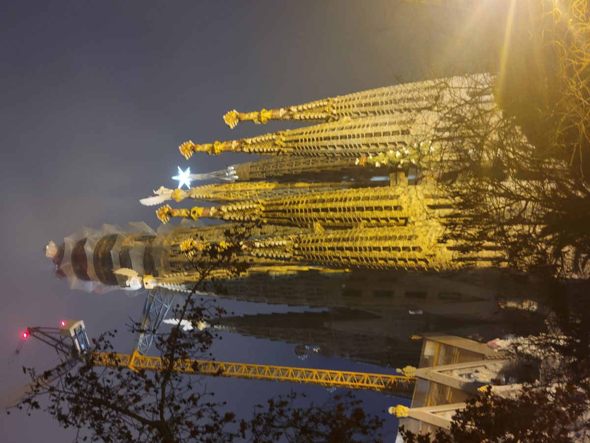 Sagrada Família illuminated at night, towers glowing gold against a dark sky with construction cranes visible
