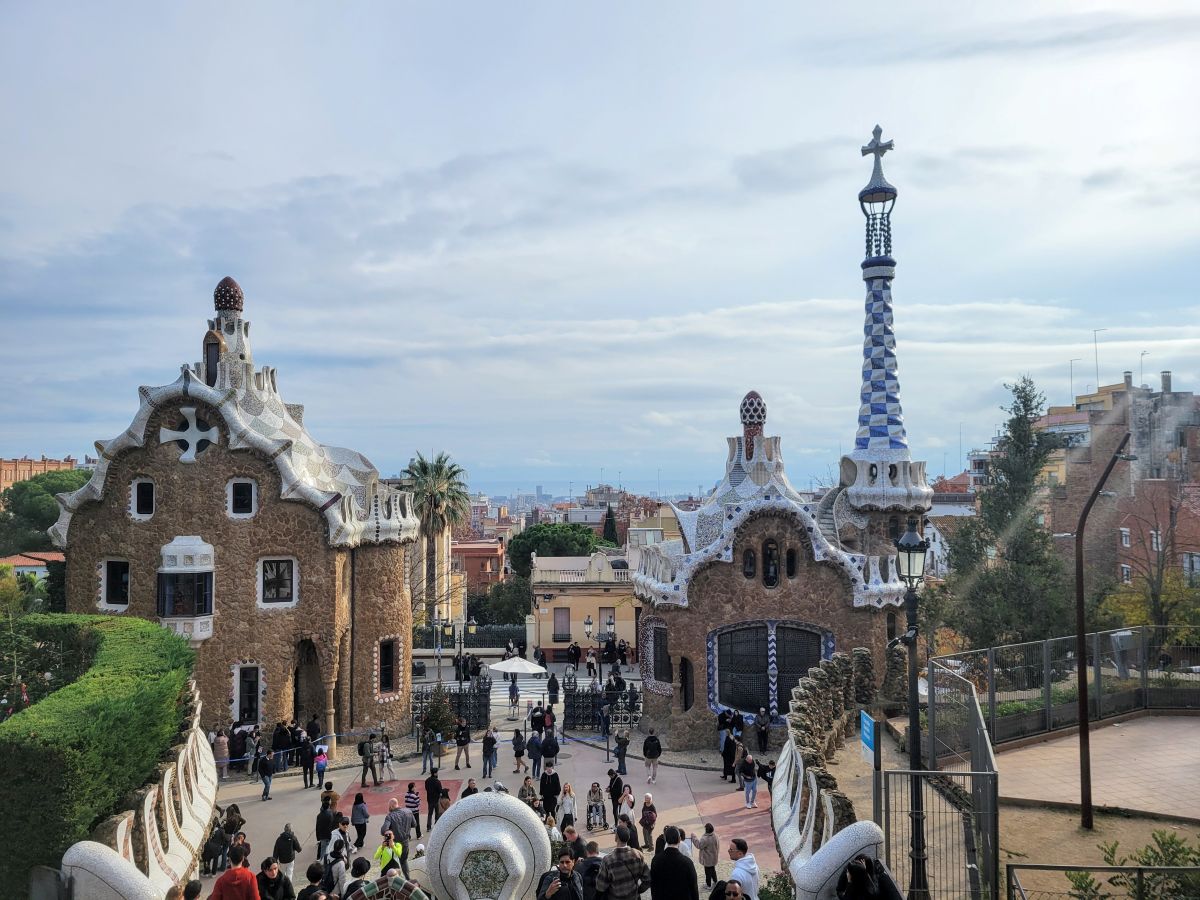 The iconic Gaudí-designed entrance pavilions at Park Güell with mosaic decorations and a tall spire, Barcelona city behind