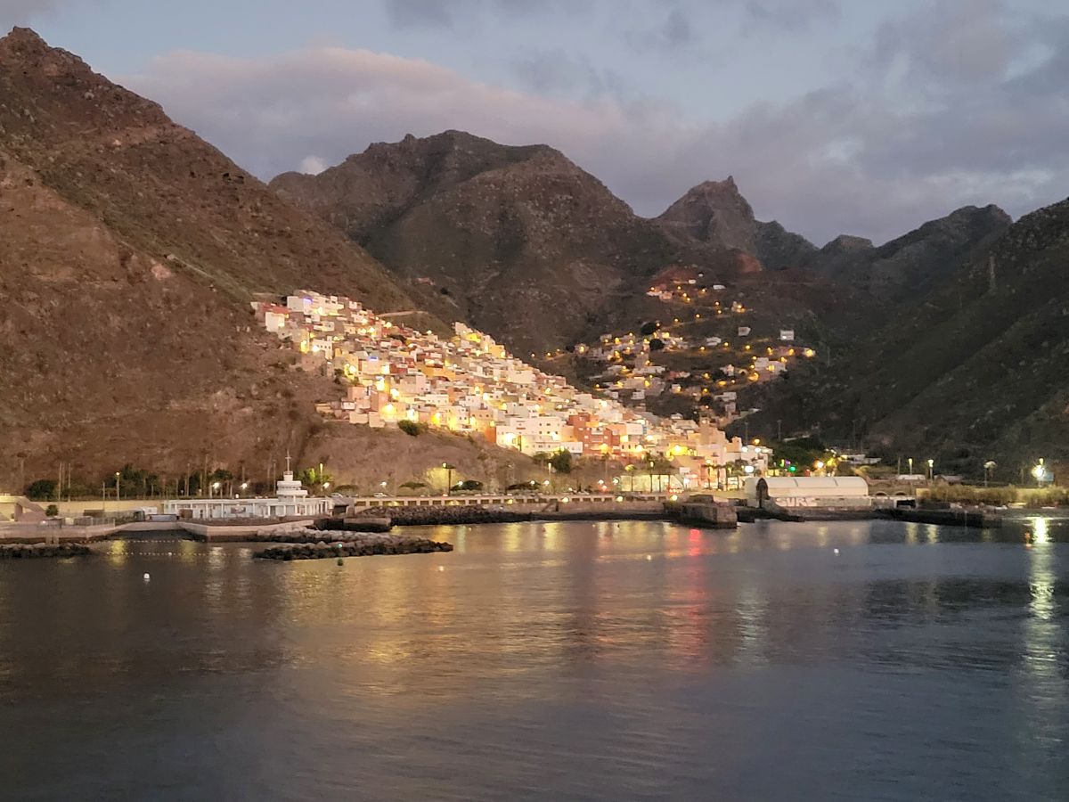 A hillside village brightly lit at night, its lights reflected in the harbor, dramatic mountains rising behind — arriving in Tenerife by ship