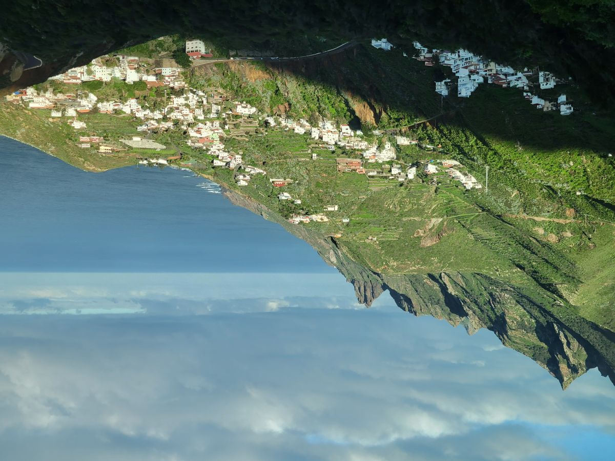 An aerial view of a white village perched on a green hillside in the Anaga Mountains, dropping steeply to the sea