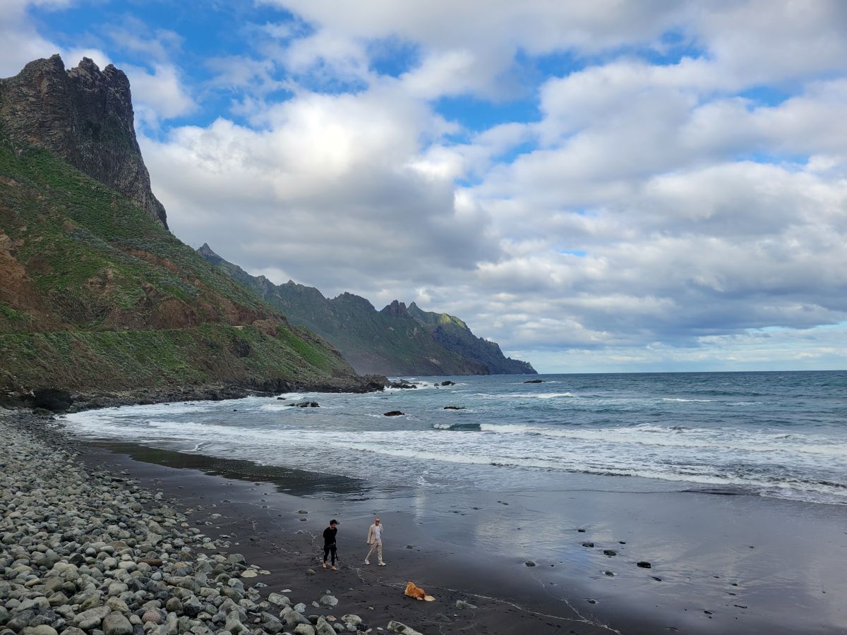 Playa del Roque de las Bodegas — a remote dark-sand beach with dramatic Anaga sea cliffs and two people walking the shore