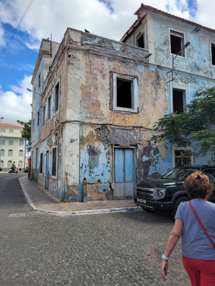 A crumbling blue-painted colonial building on a cobblestone corner in Mindelo, Cape Verde, a woman walking past
