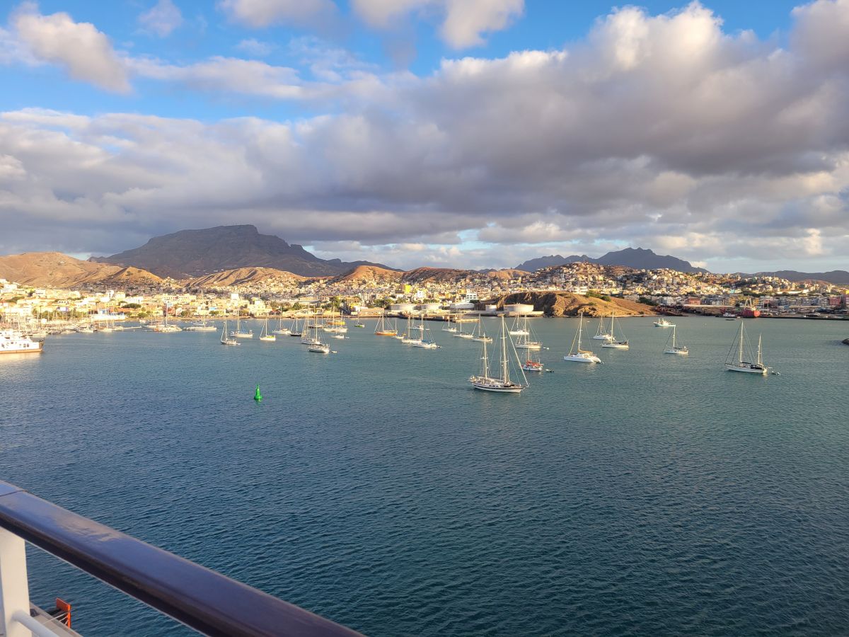 Departing Mindelo — dozens of sailboats anchored in the harbor with the colorful city and mountains of São Vicente behind, seen from the ship's deck