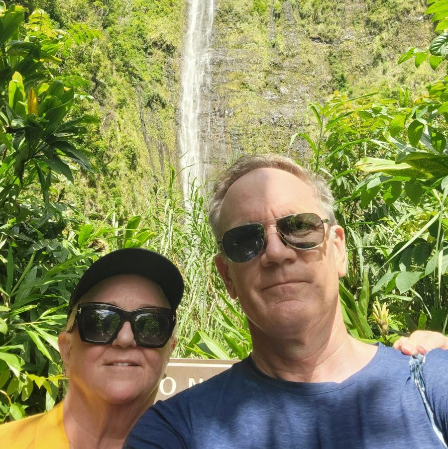 Keith and Shirley selfie in front of Waimoku Falls, a tall waterfall cascading down a lush green cliff