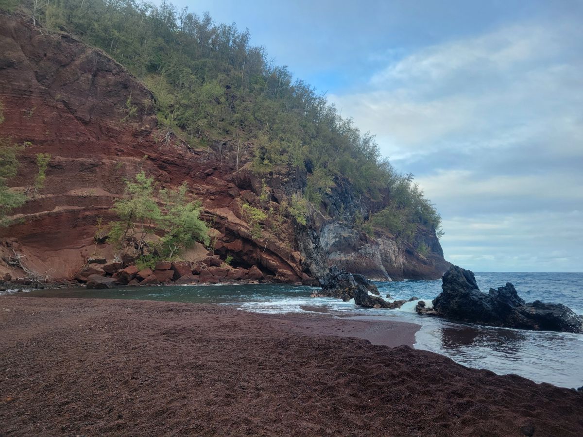 Kaihalulu Red Sand Beach — deep red volcanic cinder sand with dramatic red cliffs and turquoise water, Hana