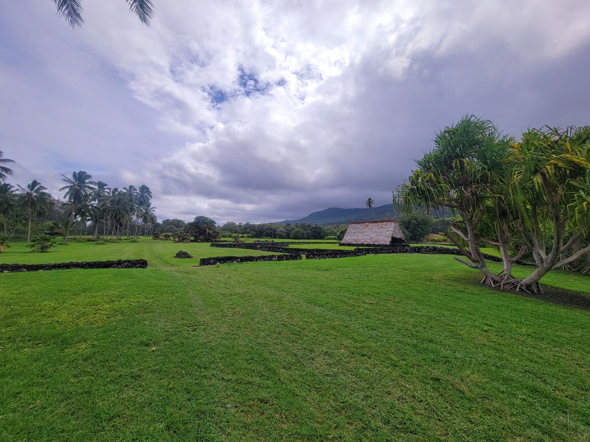 Pi'ilanihale Heiau — broad green lawn with a traditional thatched hale, lava rock walls, and palm trees at Kahanu Garden