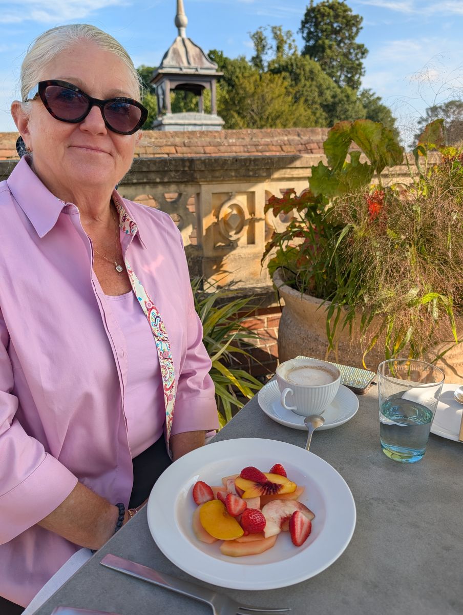 Shirley at breakfast on the terrace at Heckfield Place on the last morning, a plate of fresh fruit and a cappuccino before her, the stone garden wall and summer plants behind her