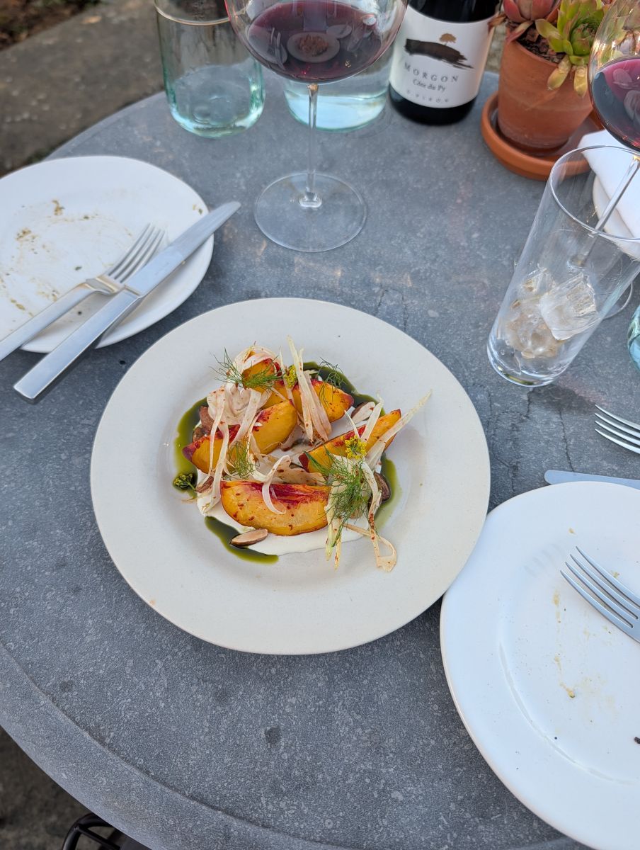 A second peach salad at Heckfield, roasted peach halves with shaved fennel and herbs over a white plate, a bottle of Morgon Beaujolais and wine glasses behind