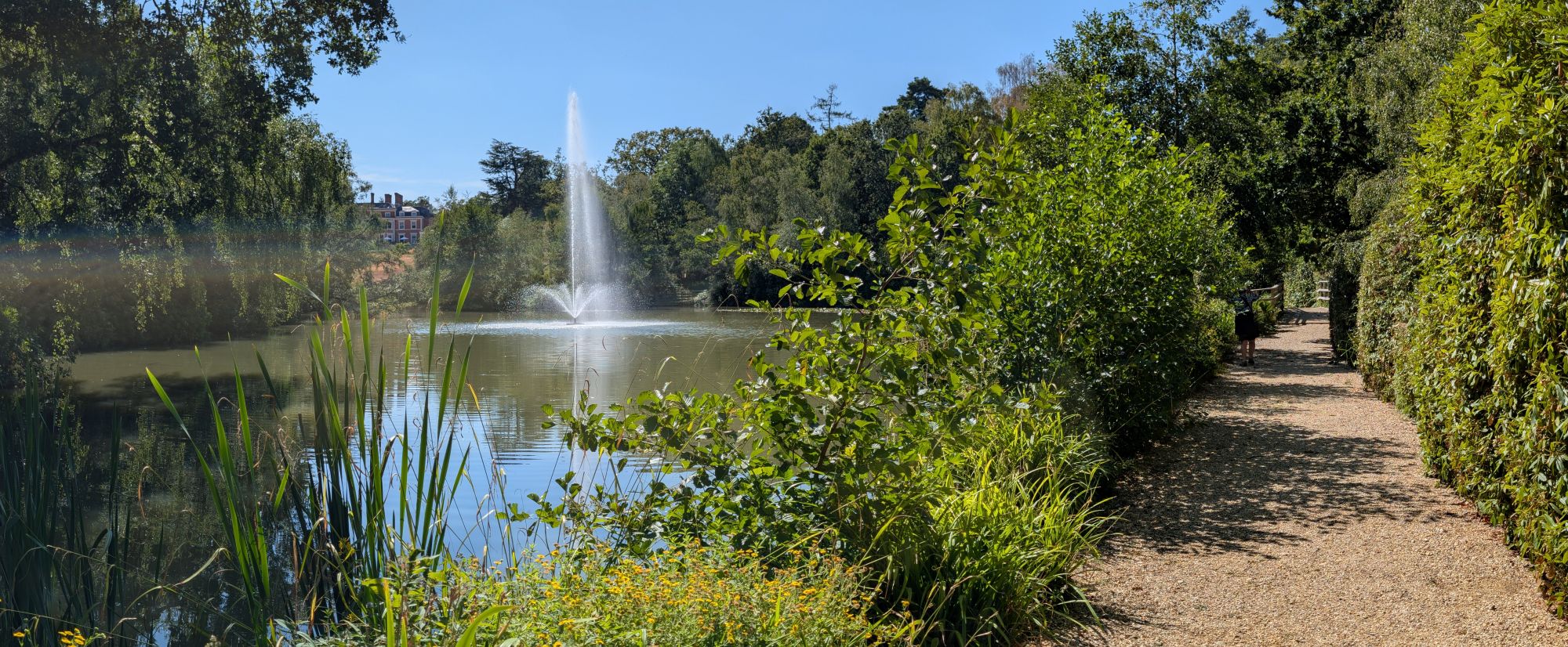 A gravel path running alongside lush hedgerows at Heckfield Place, a lake with a tall fountain beyond, the Georgian manor just visible through the trees