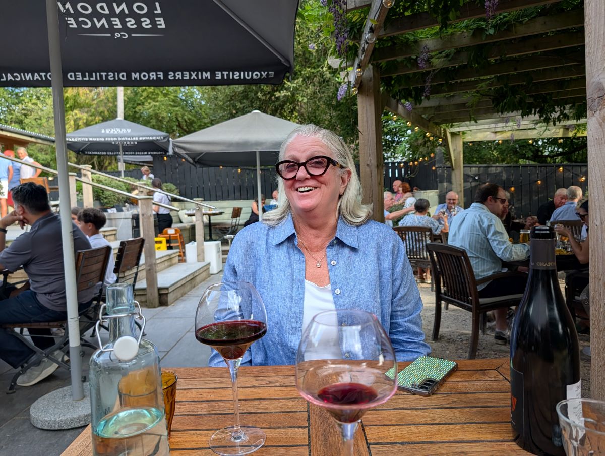 Shirley smiling at dinner at The Mutton, a local pub in Hampshire, two glasses of red wine on the wooden table, a busy summer garden terrace behind her