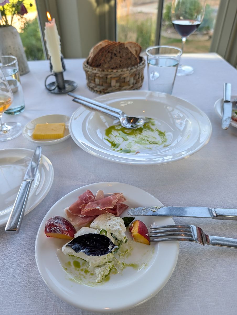 The peach and burrata salad with San Daniele ham at Heckfield Place, an empty plate smeared with green herb oil beside it, bread basket and wine glasses on the table