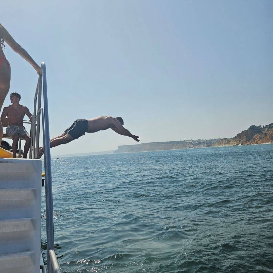 Keith diving from the upper deck of a catamaran into the Atlantic, golden limestone cliffs stretching along the coast behind him