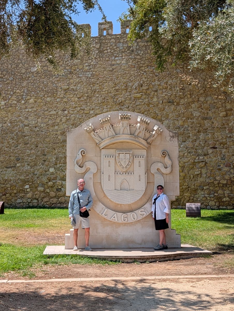 Keith and Shirley standing on either side of the large Lagos coat of arms carved in stone, the old city walls and olive trees behind them
