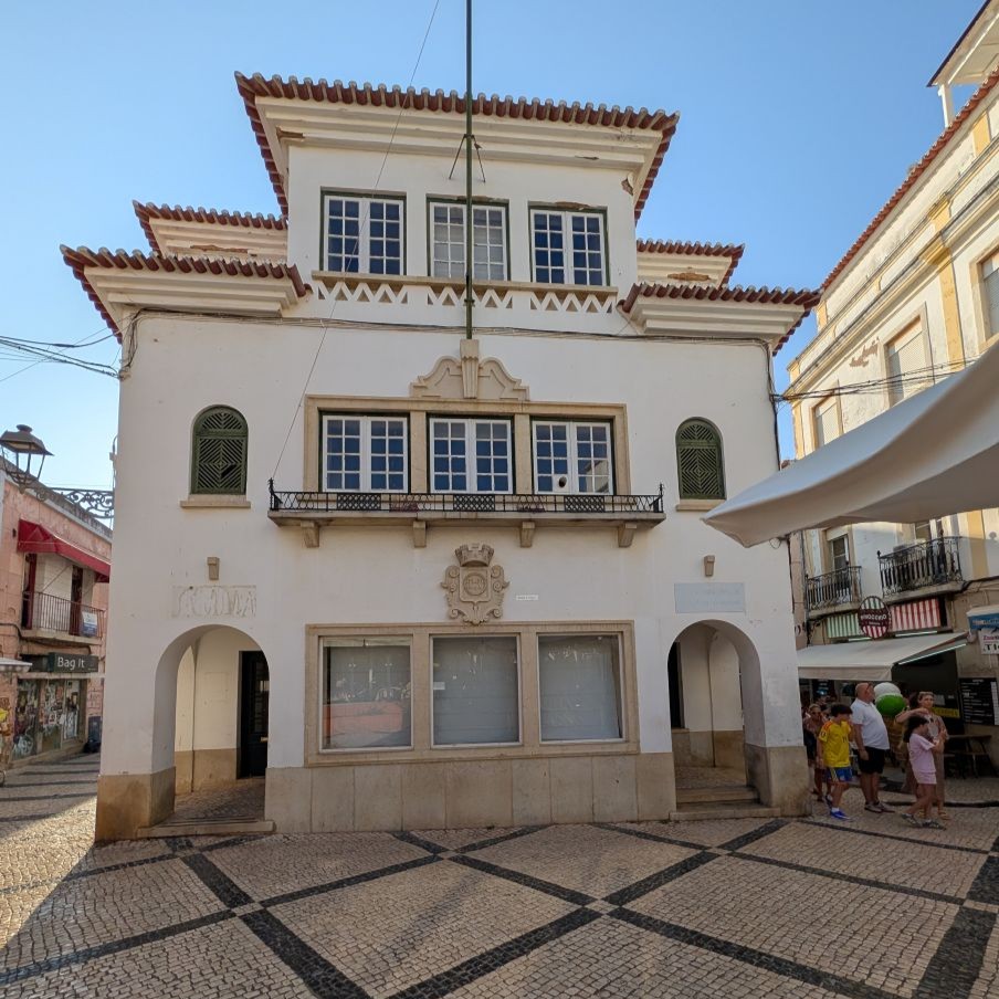 A white art deco building with green shutters and an arched entrance facing a cobblestone square in the old town of Lagos — a vacant storefront with an apartment above