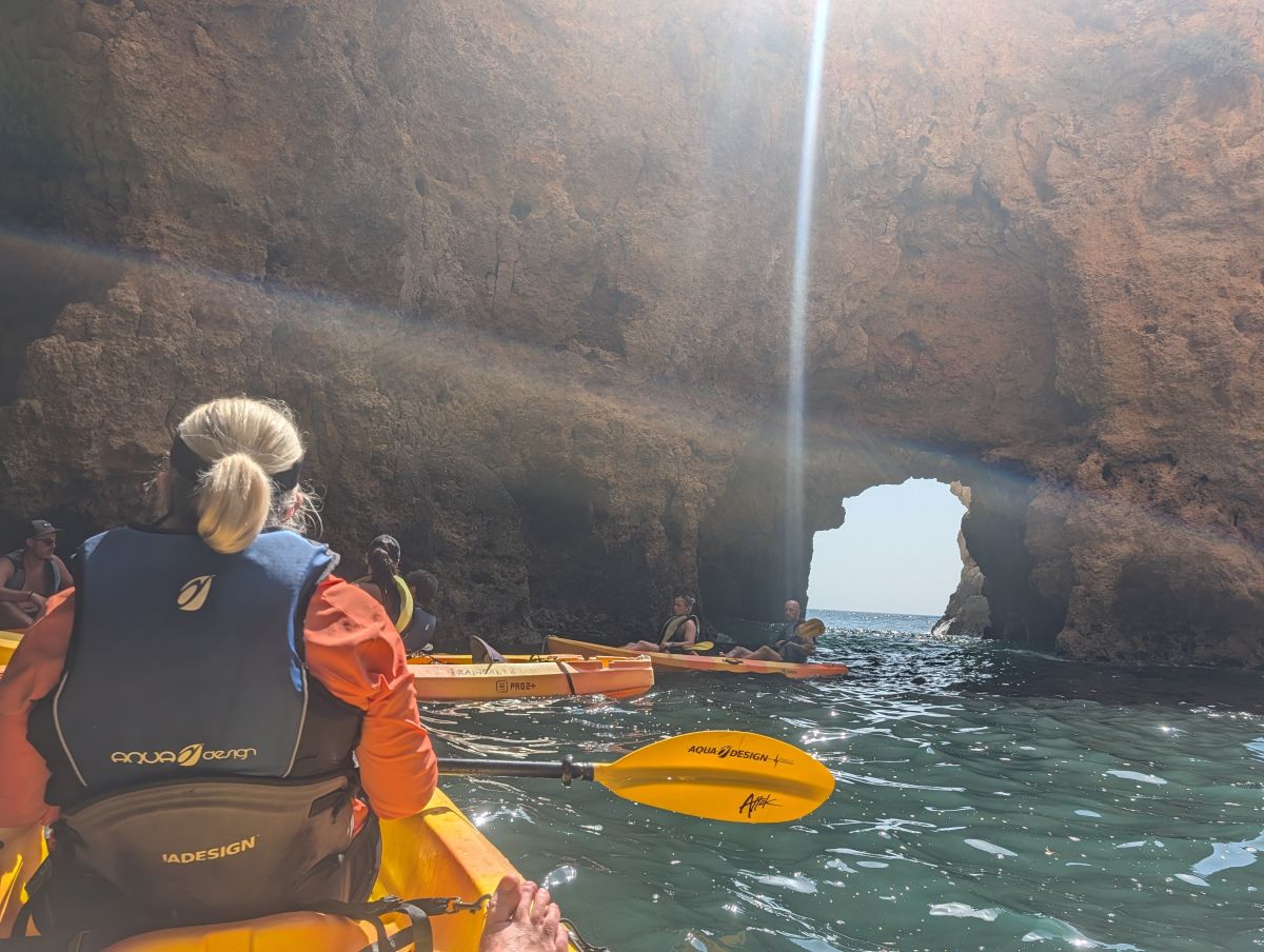 Kayakers inside a dramatic sea cave at Ponta da Piedade, a beam of light streaming through an opening in the rock above, the arch framing the open ocean beyond