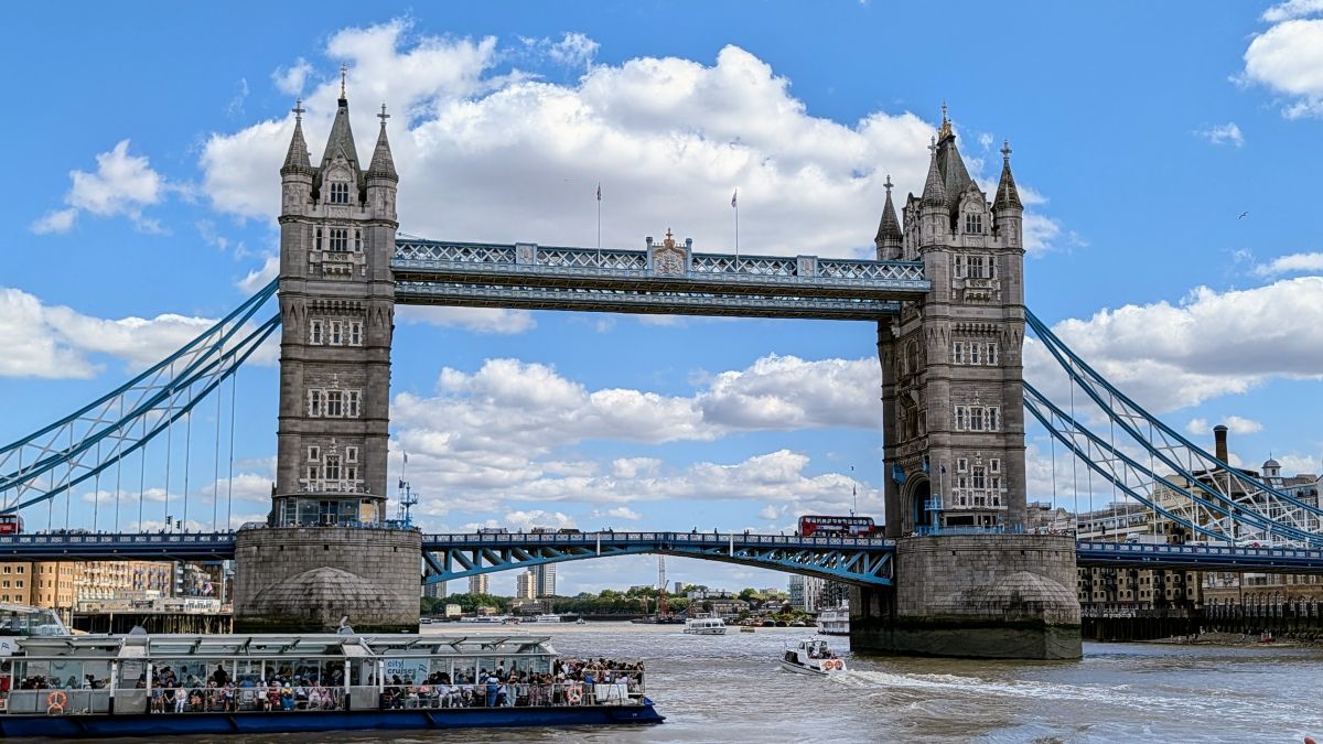 Tower Bridge over the Thames on a sunny day, a City Cruises boat full of tourists passing beneath, a red double-decker bus crossing above