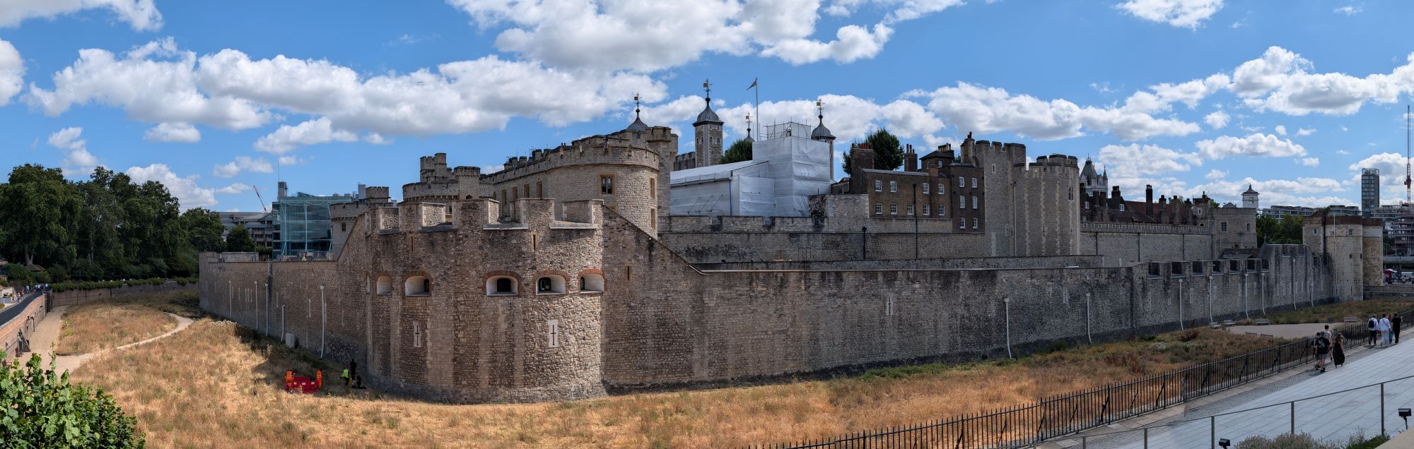 Panoramic view of the Tower of London, its stone walls and towers stretching the width of the frame, dry summer grass in the moat below
