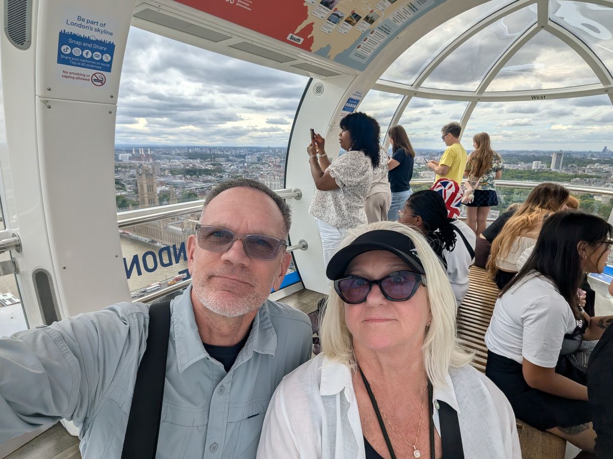 Keith and Shirley selfie inside a London Eye capsule, the city stretching below, Westminster visible through the glass, other visitors looking out behind them