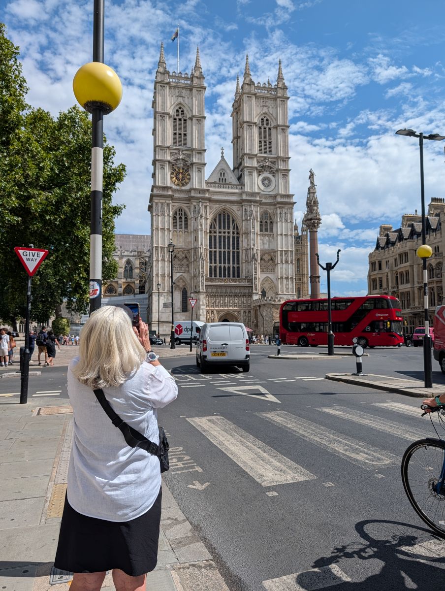Shirley photographing Westminster Abbey from across the street, a red double-decker bus passing, the Abbey's Gothic towers filling the sky