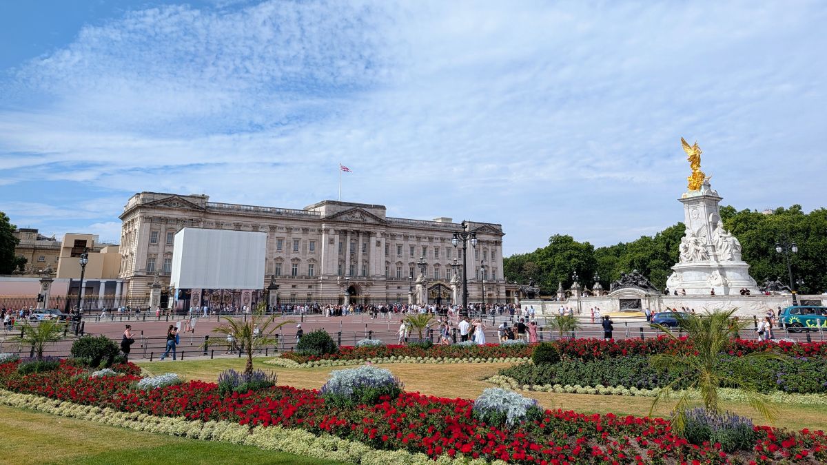 Buckingham Palace in sunshine, red flowers and the Victoria Memorial in the foreground, crowds gathered at the gates